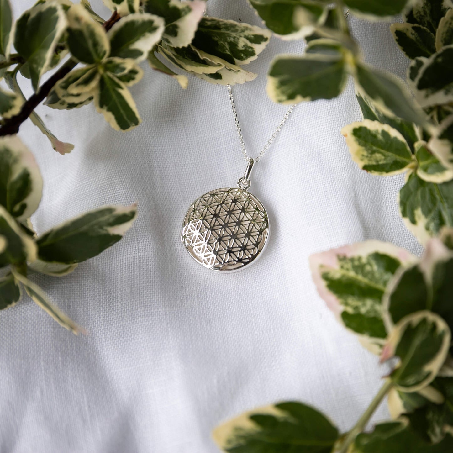 Silver geometric photo locket necklace on a white fabric background with green leaves.
