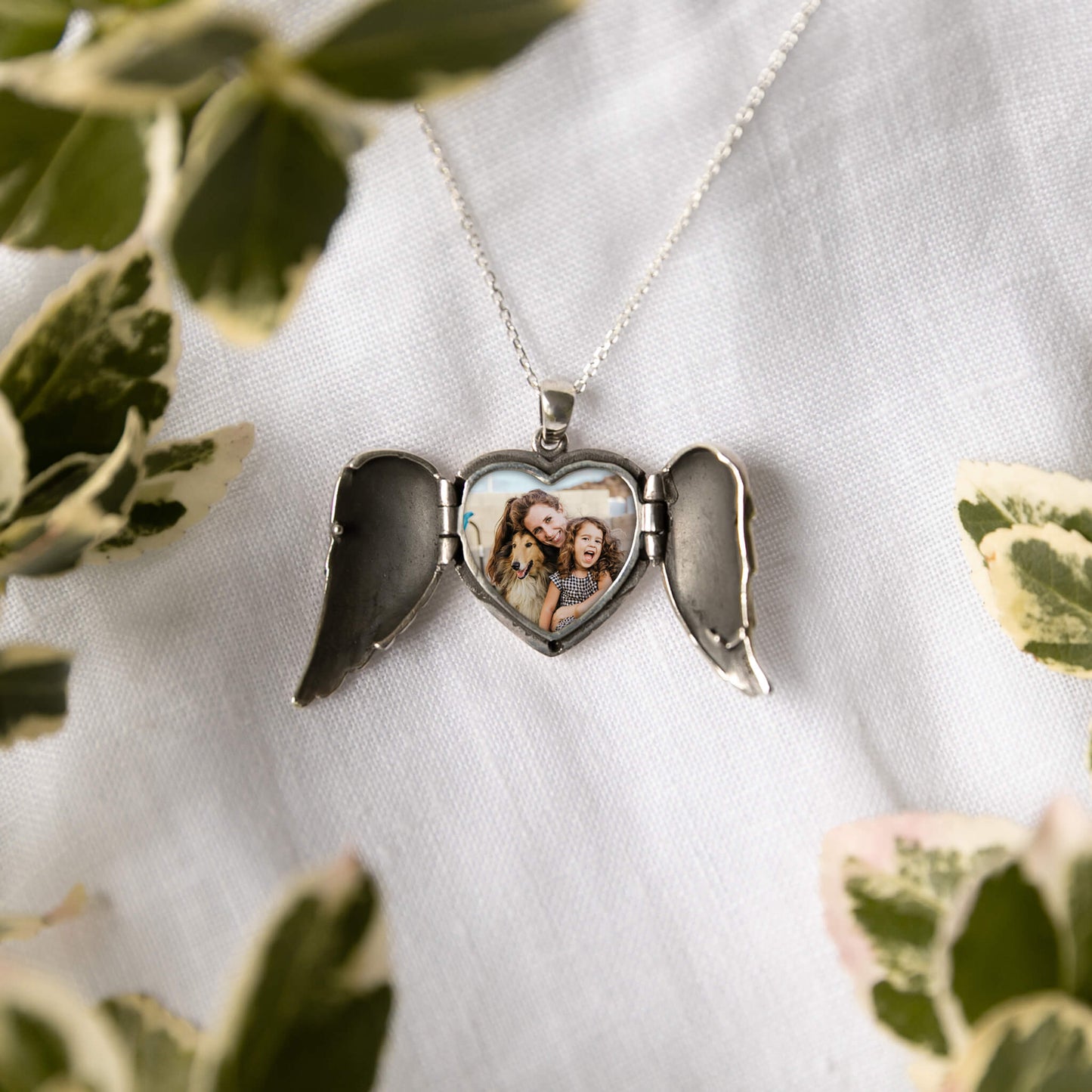 Heart-shaped locket with angel wings on a white fabric background with green leaves.