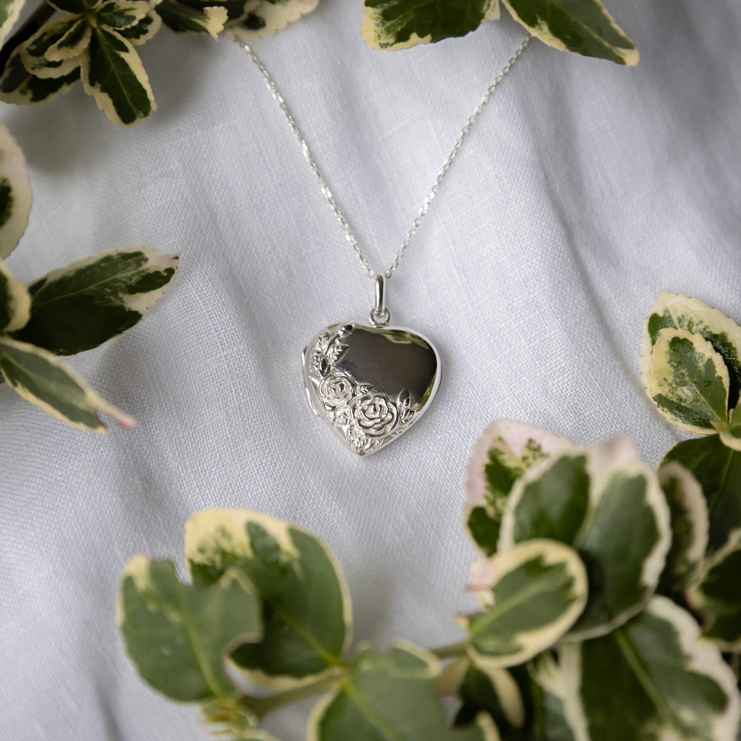 Silver heart-shaped pendant necklace on a white fabric background with green leaves.