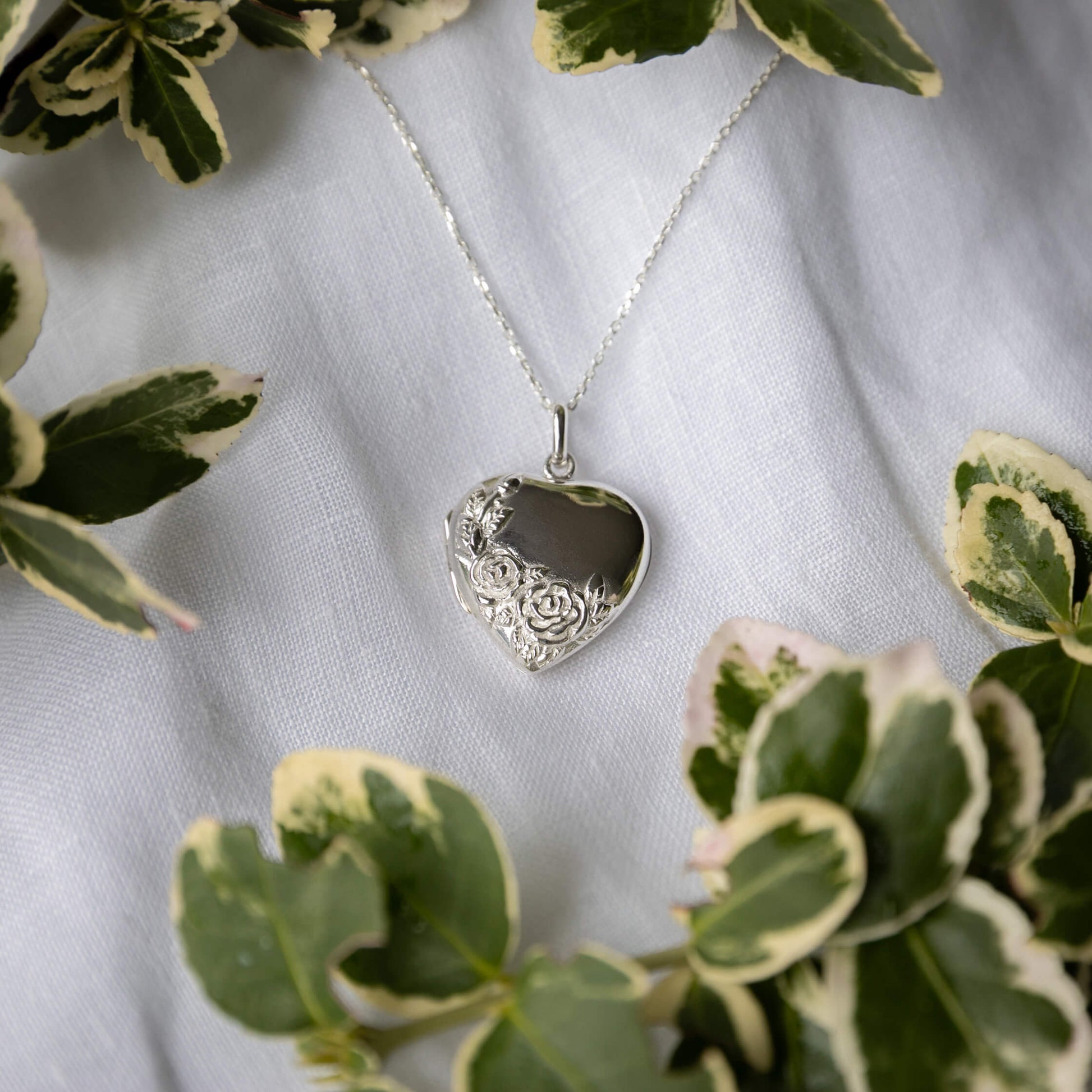 Silver heart-shaped pendant necklace on a white fabric background with green leaves.