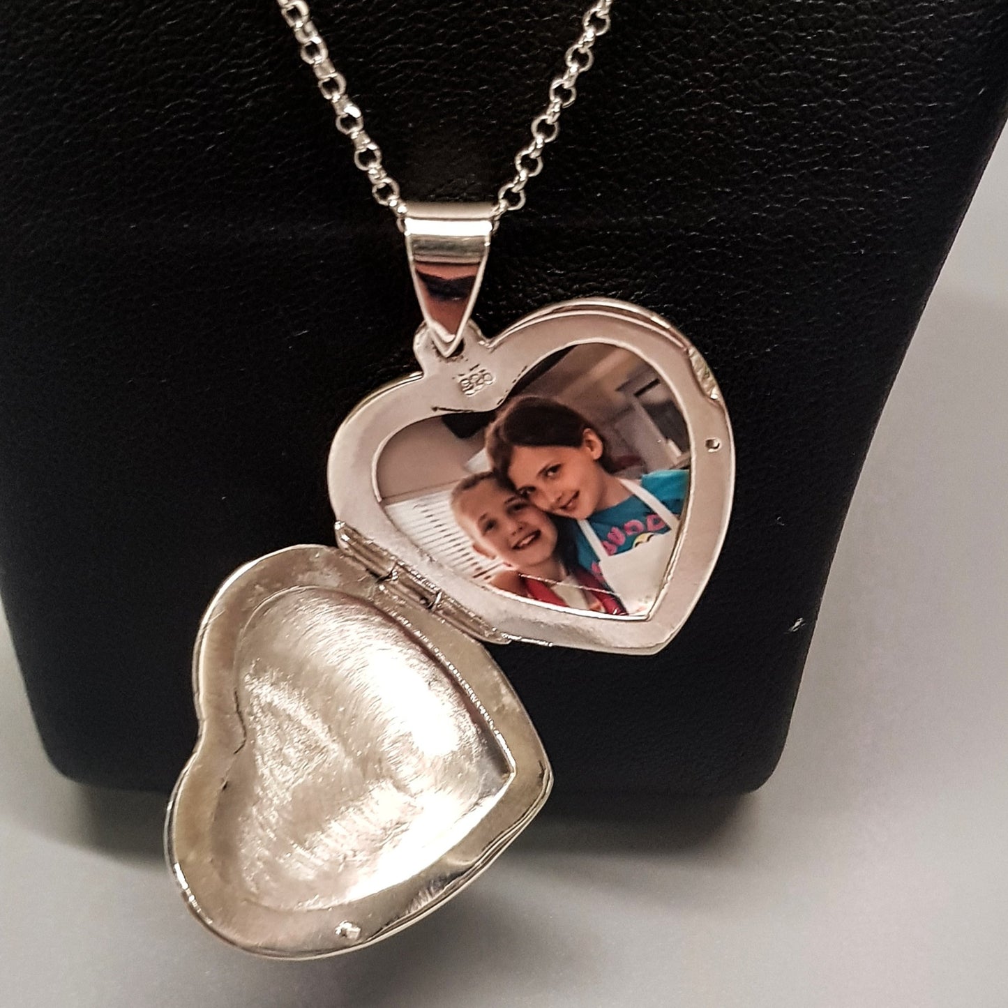 Large silver heart shaped photo locket necklace on silver chain displayed on a black bust. Locket is open showing photo of two little girls inside.