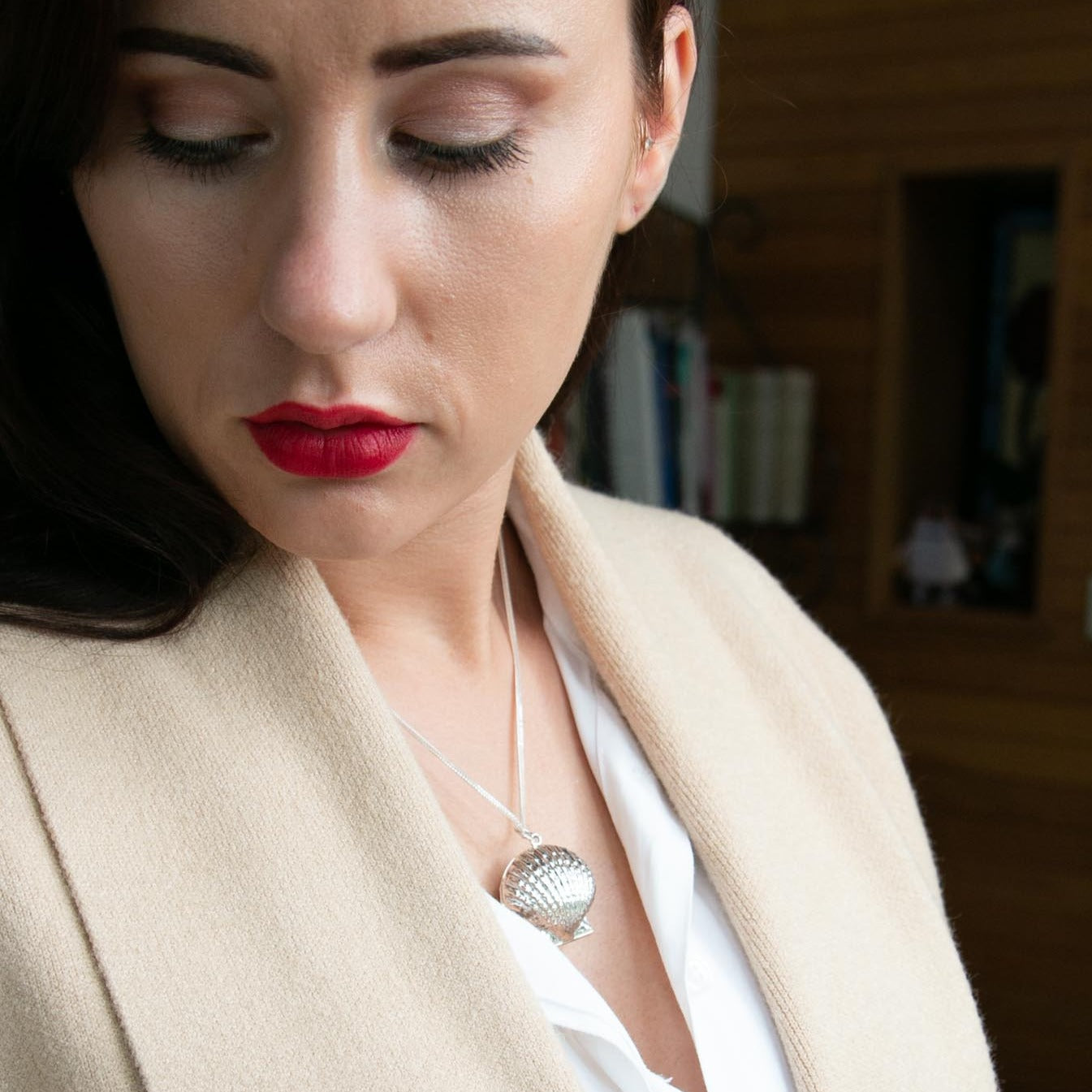 Model with red lips, wearing beige coat and white shirt and sterling silver clam shell-shaped photo locket pendant. Model is looking downwards.