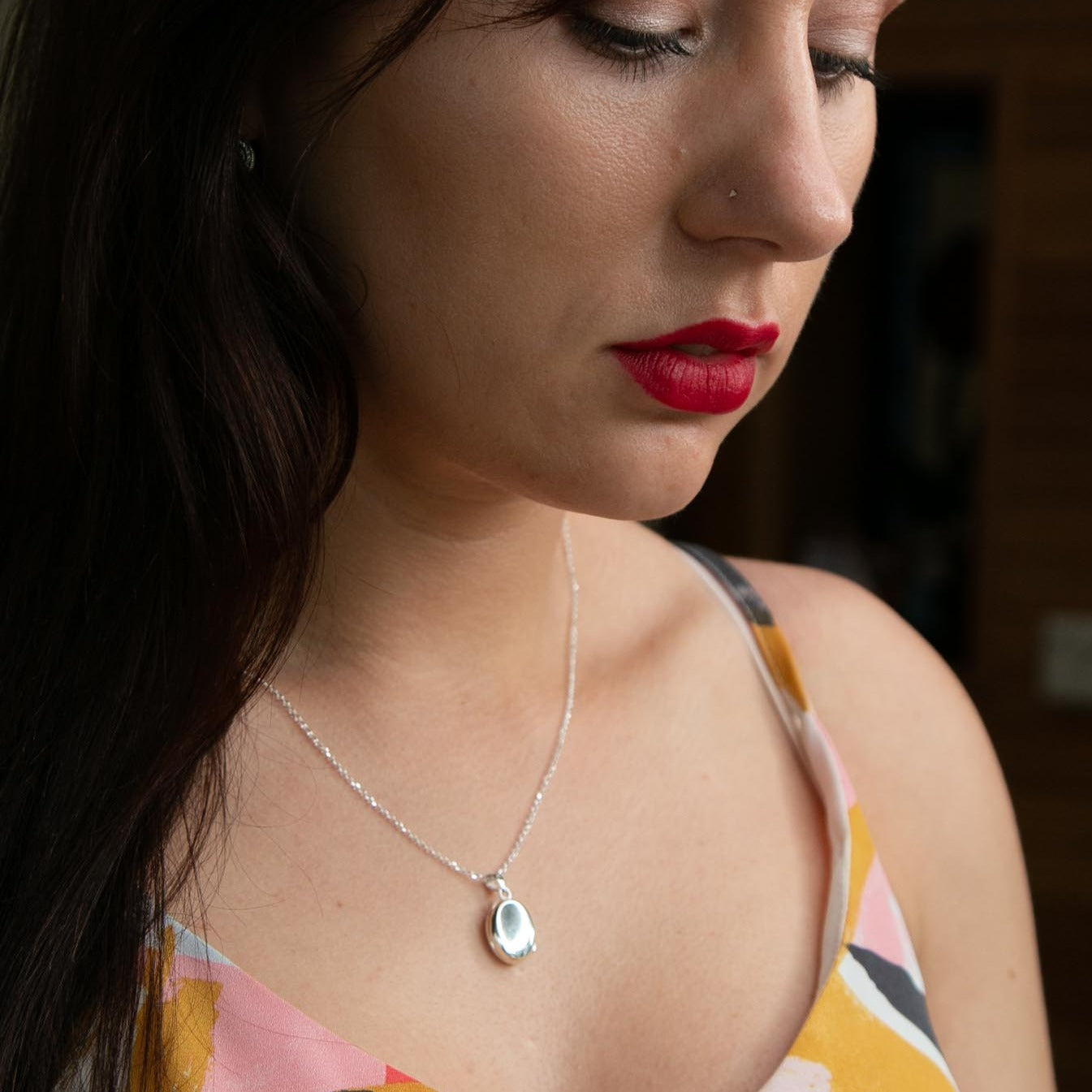 Model wearing small silver locket necklace on silver chain. Model is looking down and wearing colourful top.